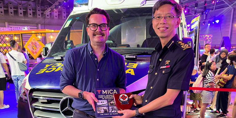 51 Medical Director Associate Professor David Anderson presenting the Mobile Intensive Care 51 (MICA) book to SCDF Director of Emergency Medical Services Department Senior Assistant Commissioner Yong Meng Wah in Singapore in 2023. Both men were standing in front of an ambulance in an exhibition hall.
